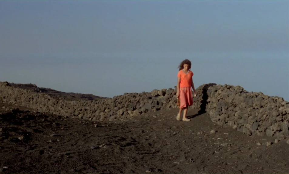 Casa De Lava (1994) movie still: wide — A volcanic landscape with black, rocky soil and a low, dry-stone wall. A woman walks slowly along the path…