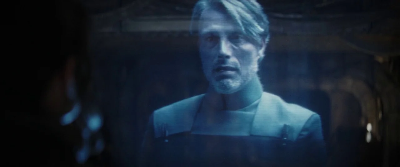 Rogue One: A Star Wars Story (2016) movie still: medium — A man appears as a translucent, flickering blue hologram within a dark interior space. The…
