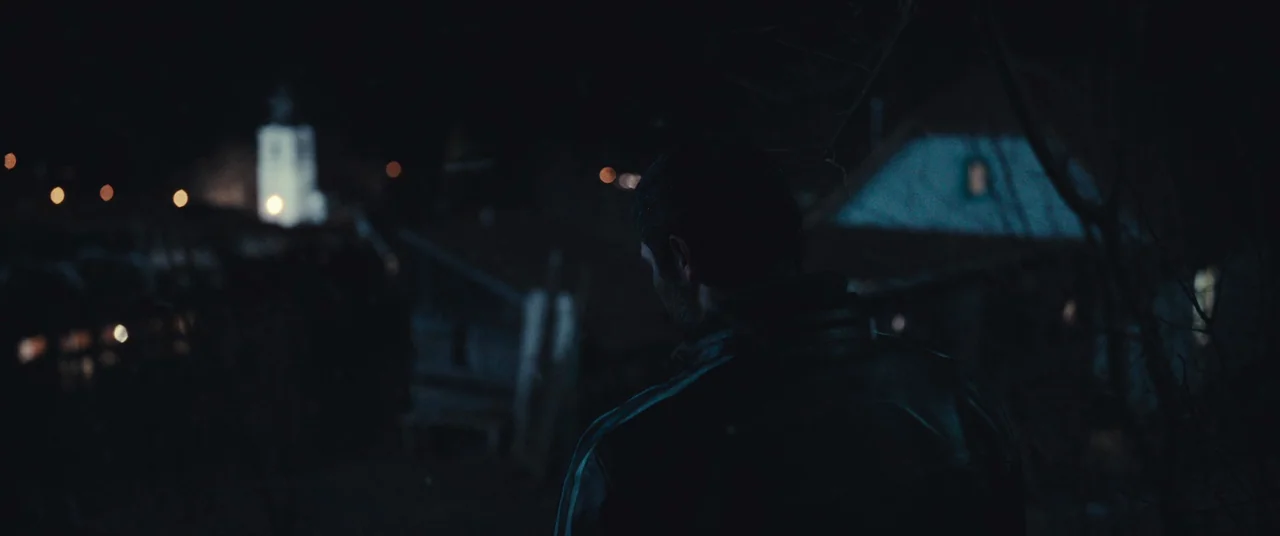 R.M.N. (2022) movie still: medium — A man stands in the dark outdoors overlooking a cluster of houses at night. A bright illuminated tower stands in…