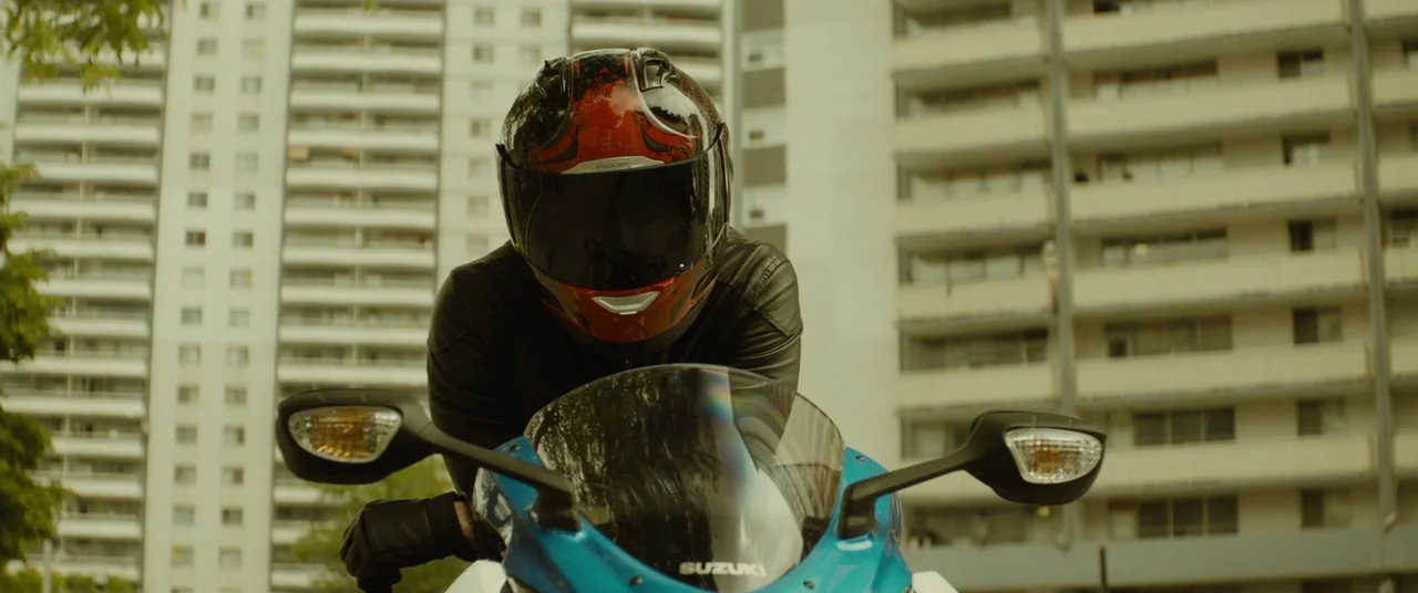 Enemy (2013) movie still: medium — A motorcycle rider wearing a red and black helmet sits on a blue sports bike. High-rise apartment buildings stand…