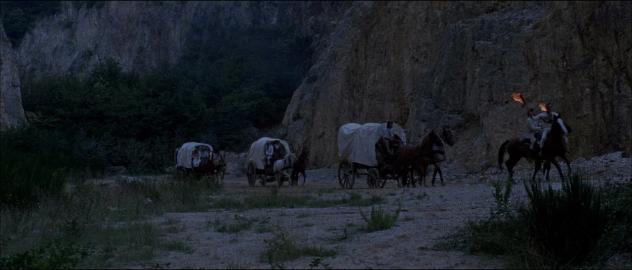 The Grand Duel (1972) movie still: wide — A group of covered wagons and horsemen travel through a rocky, narrow canyon floor. The riders lead the…