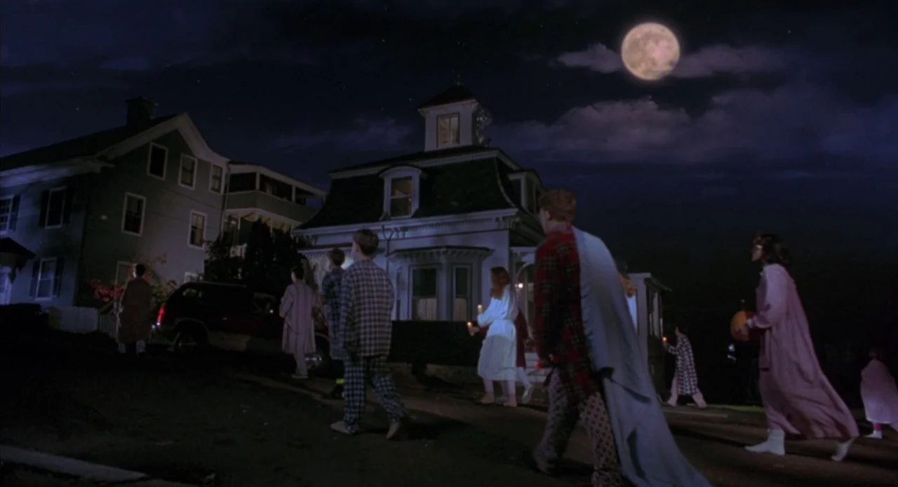 Hocus Pocus (1993) movie still: wide — A quiet residential street at night featuring dark houses and a large full moon. A group of people dressed in…