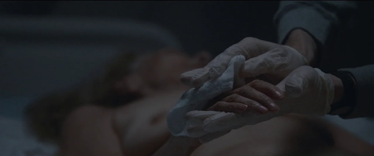 The Good Nurse (2022) movie still: closeup — A dimly lit room contains a body resting on a metal examination table. Hands wearing white latex gloves…