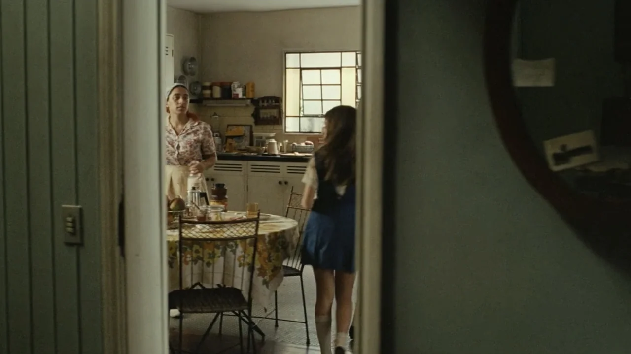 I'm Still Here (2024) movie still: medium — A 1970s-style domestic kitchen features a round table with a yellow floral tablecloth. A woman stands…