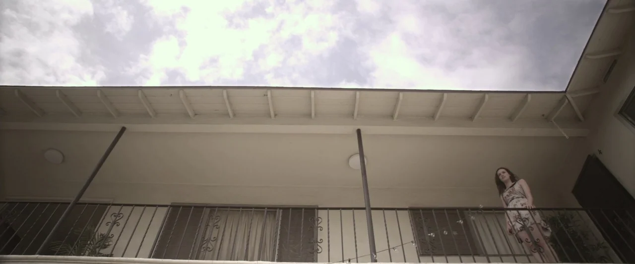 Starry Eyes (2014) movie still: wide — A two-story stucco apartment building has a walkway with a decorative metal railing. A young woman stands on…