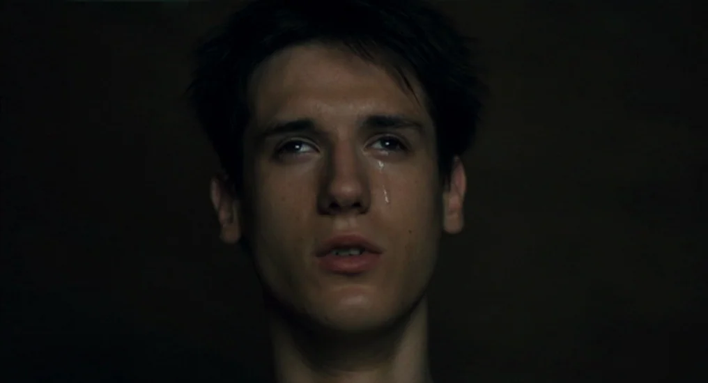 Paris, Je T’Aime (2006) movie still: closeup — A person is in a dark, nondescript room with no visible walls or furniture. A young man gazes upward…
