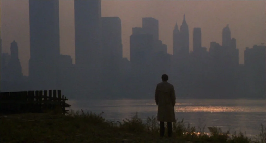 The Killing Fields (1984) movie still: wide — A murky, hazy twilight view of the New York City skyline across a body of water. A lone man stands in…