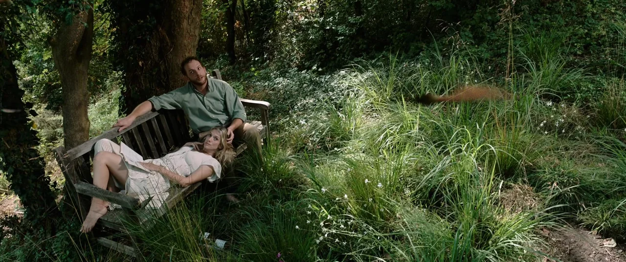 Oxygen (2021) movie still: wide — A wooden bench sits amidst tall grass and weeds in a shaded forest clearing. A man and a woman rest together on…