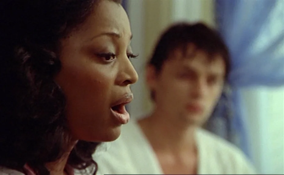 Diva (1981) movie still: closeup — A close-up view of a woman speaking while a man is out of focus behind. A blue sheer curtain hangs in the window…