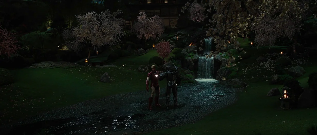 Iron Man 2 (2010) movie still: wide — A manicured night landscape features cherry blossom trees and a small stone waterfall. Two armored figures…