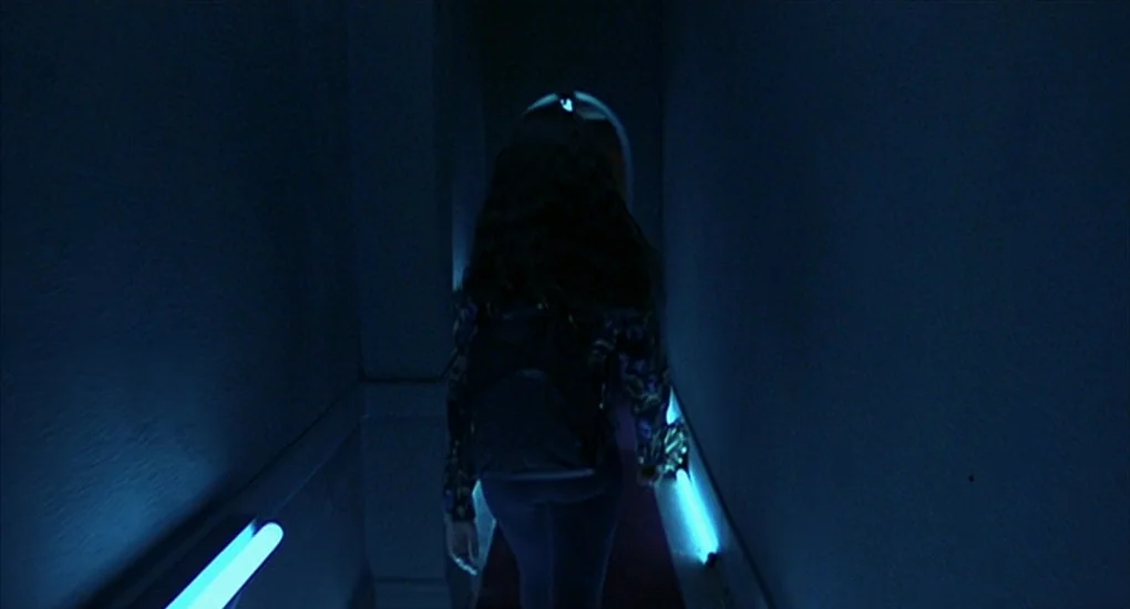 Exotica (1994) movie still: medium — A dark, narrow indoor passageway features bright blue horizontal neon light strips. A young person with long…