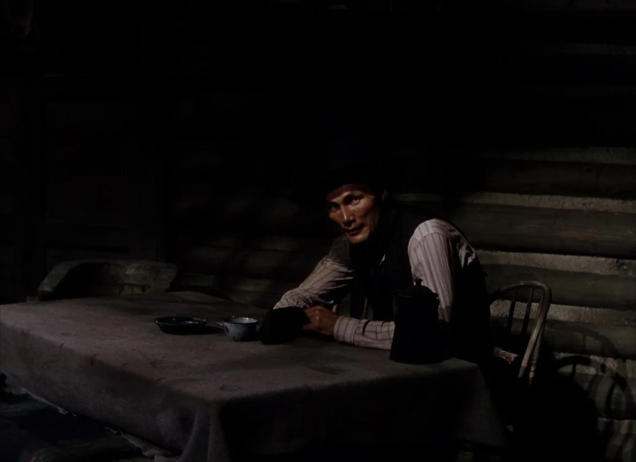 Shane (1953) movie still: medium — A dimly lit log cabin interior is defined by wooden walls and simple furniture. A solitary man sits at a table…