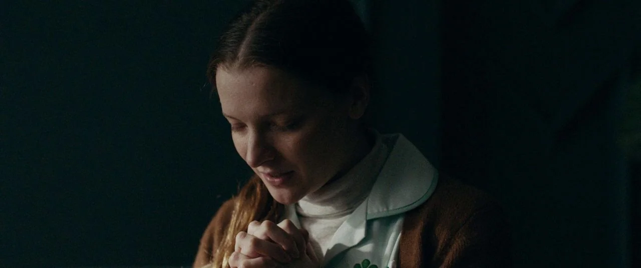 Saint Maud (2019) movie still: closeup — A dimly lit room with dark walls and soft, directional light across the subject. A young woman with her…