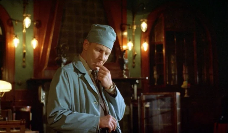 The Turn of the Century (2001) movie still: medium — An dimly lit interior study with wood-paneled walls and wall-mounted brass lamps. A man wearing…