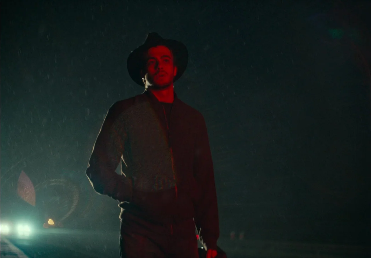 EO (2022) movie still: medium — A dark night scene on a wet highway with blurred lights in the distance. A man wearing a fedora walks through the…