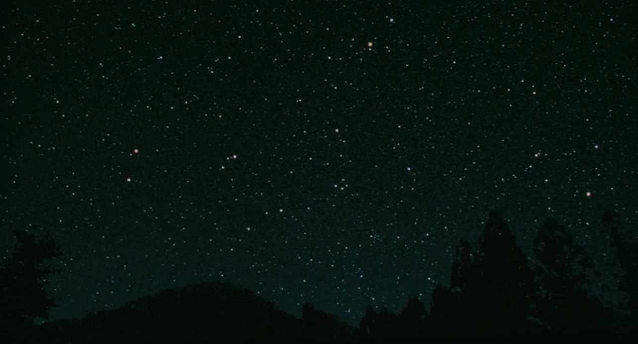 Uzumaki (2000) movie still: establishing — A dark, clear night sky is filled with numerous dense clusters of bright stars. Silhouettes of a mountain…