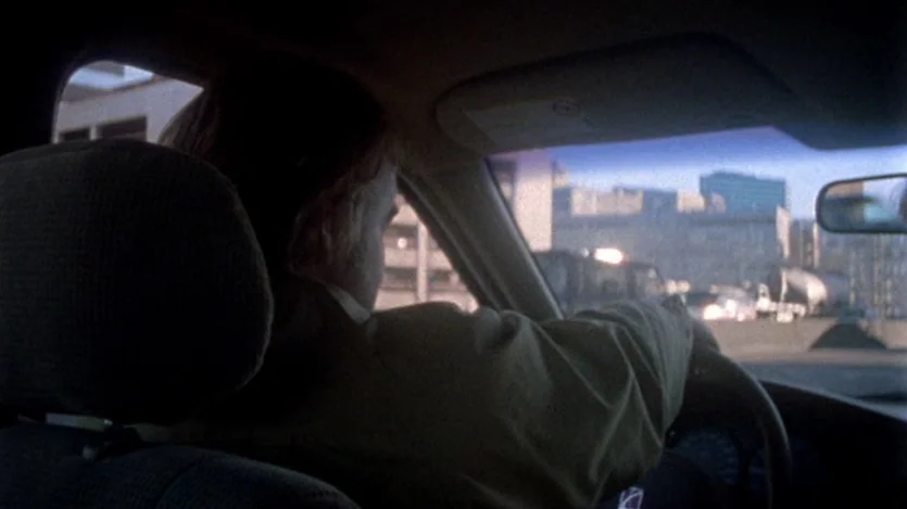 Love Liza (2002) movie still: medium — The interior of a sedan during daylight with city buildings visible through the windshield. A man sits behind…