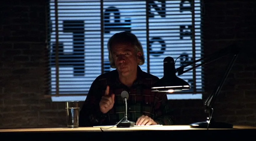 Gray’s Anatomy (1996) movie still: medium — A dark office at night features a man seated before a closed window blind. He sits behind a desk with a…