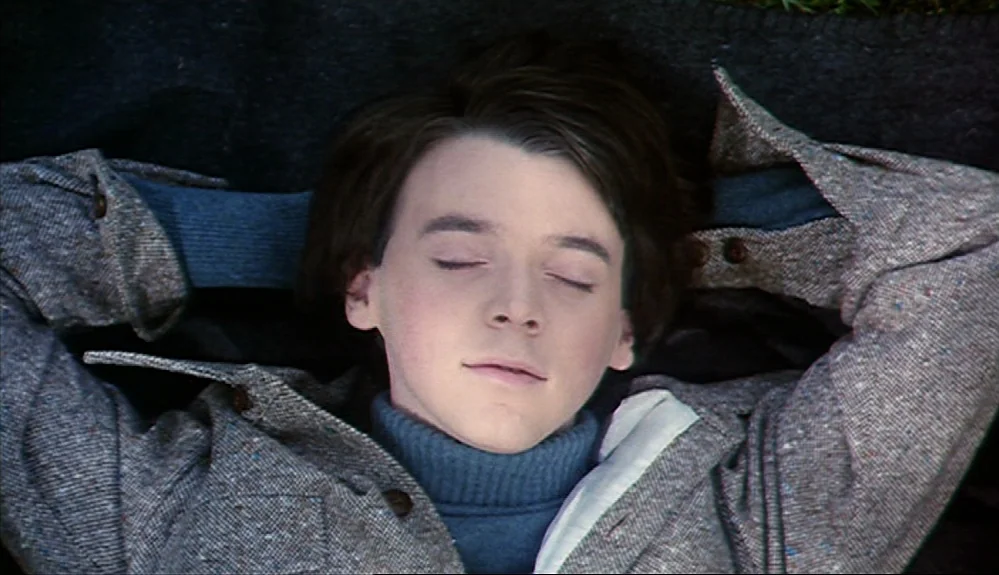 Harold and Maude (1971) movie still: closeup — A teenage boy lies flat on his back against a dark surface outdoors. He keeps his eyes closed with…