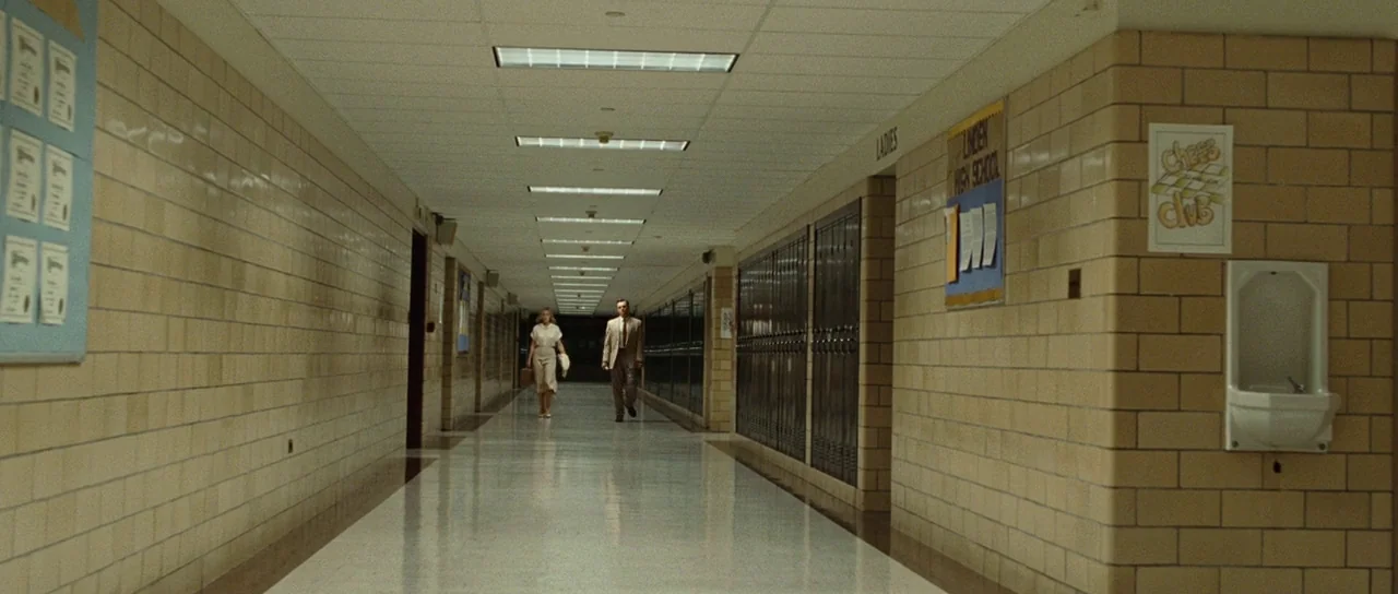 Revolutionary Road (2008) movie still: wide — A long school corridor features beige brick walls and rows of recessed metal lockers. A man and a…