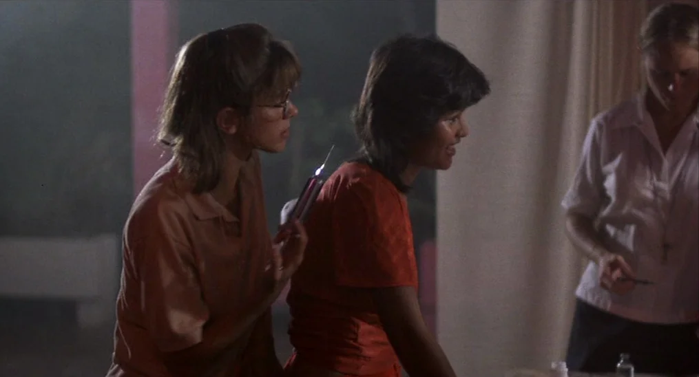 Salvador (1986) movie still: medium — A dimly lit room features two women interacting near a workstation with medical supplies. One woman holds a…