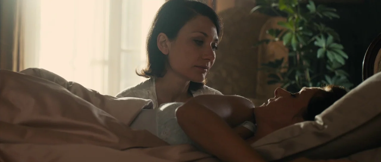 The Duke of Burgundy (2014) movie still: medium — A well-lit bedroom features a woman sitting over someone resting under beige bedding. A large…