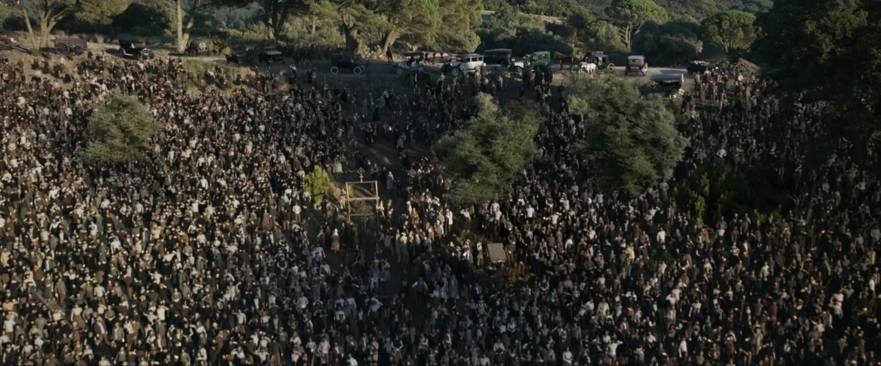 Fatima (2020) movie still: establishing — A vast, sloped grassy hillside is densely populated by a large crowd of people. Numerous vintage…