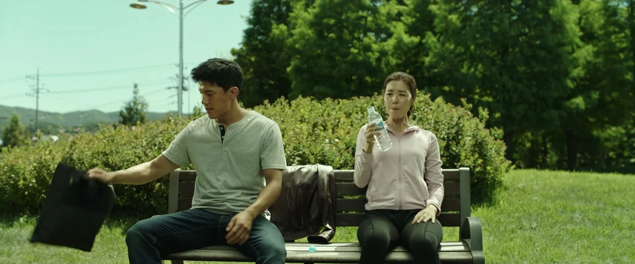 The Gangster, the Cop, the Devil (2019) movie still: medium — An outdoor park setting with lush green trees and a wooden bench. A man holding a…