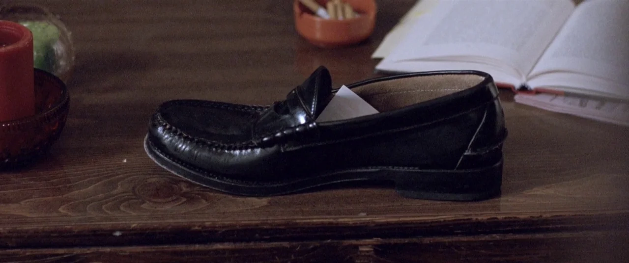The End of the Tour (2015) movie still: closeup — A worn wooden table features a black leather loafer resting in the center. An open book and a…