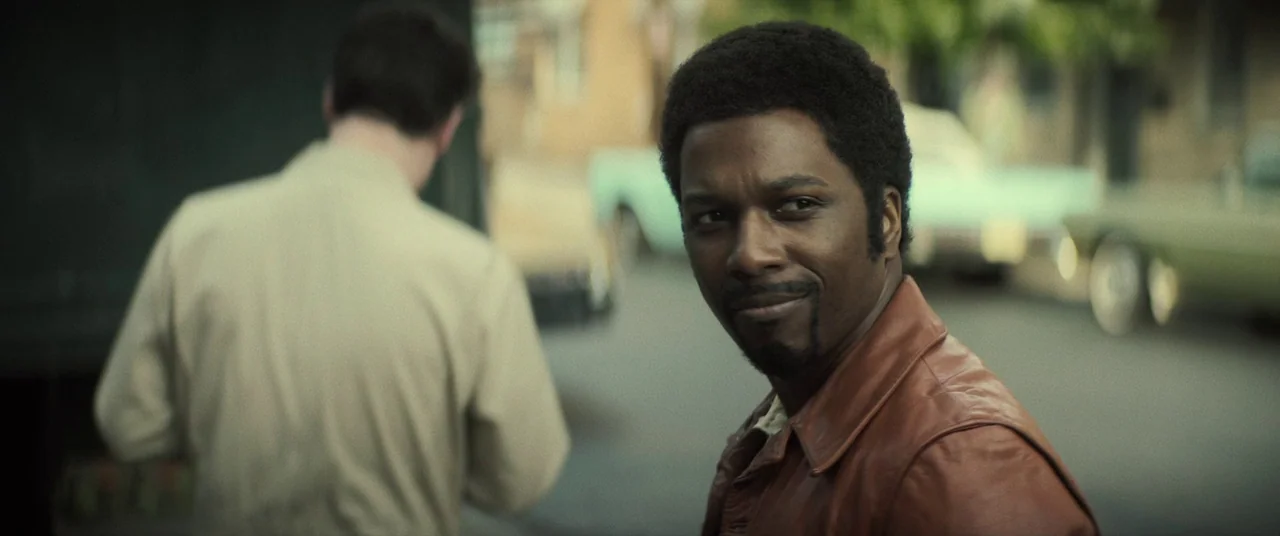 The Many Saints of Newark (2021) movie still: medium — A residential city street features parked vintage cars in the blurred background. A black man…