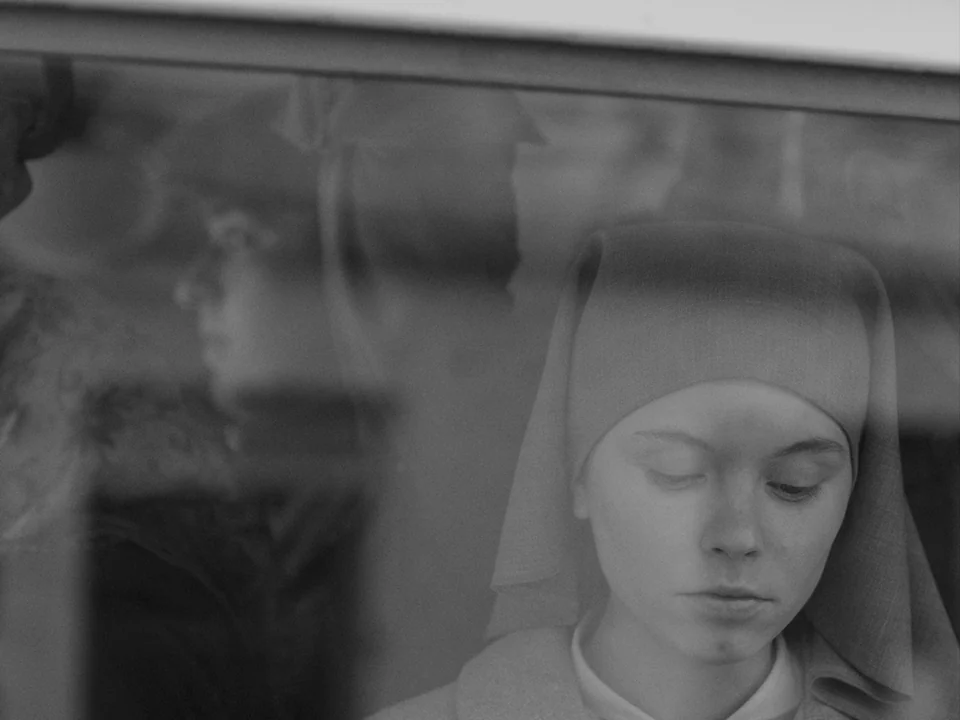 Ida (2013) movie still: closeup — A nun sits behind a reflective window pane on a moving vehicle. She looks downward while blurred shapes of other…
