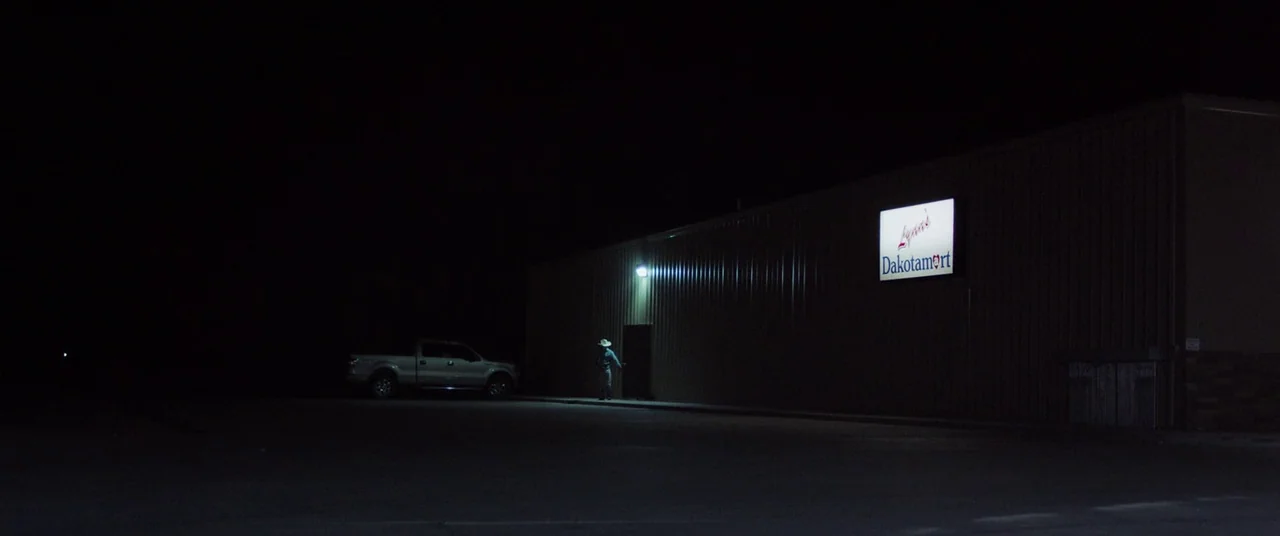 The Rider (2017) movie still: wide — A large commercial building stands in darkness at night with a single lit sign. A lone figure walks toward a…