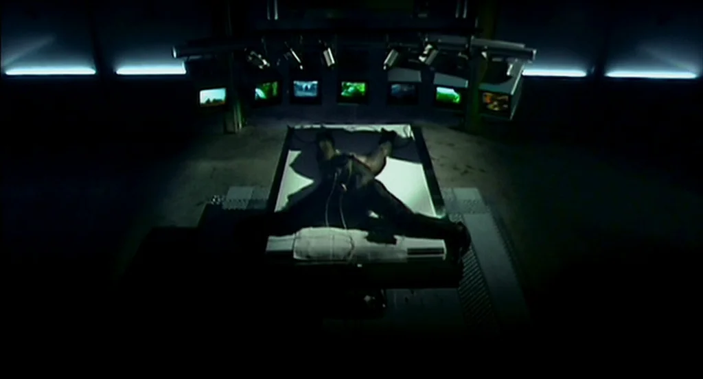 28 Days Later (2002) movie still: wide — A dark, industrial surveillance room contains a single white metal examination table in center. A man lies…