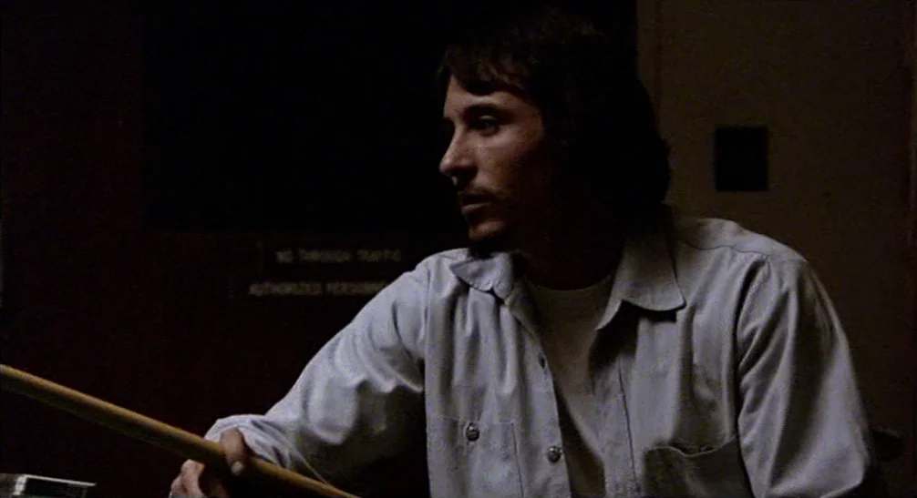 Coming Home (1978) movie still: medium — A dark, dimly lit room contains a person holding a wooden object. The man in a light shirt looks away from…