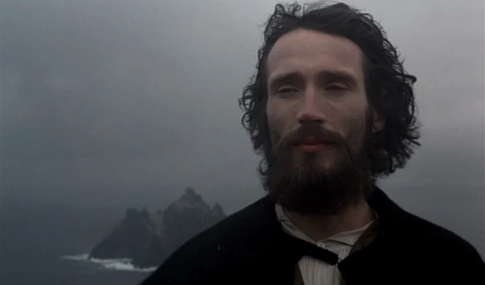 Heart of Glass (1976) movie still: closeup — A man stands before a dark, craggy island formation rising from a grey sea. The sky is completely…