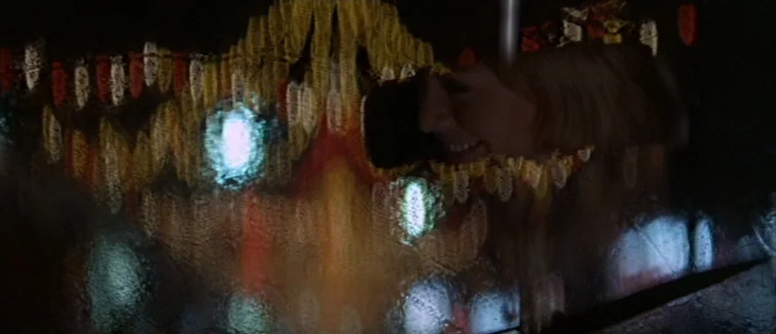 Images (1972) movie still: closeup — A night scene shows a woman peering through a rain-covered window pane. Multicolored neon lights from the city…