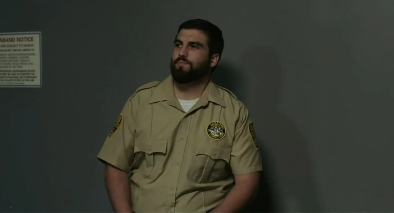 Elles (2018) movie still: medium — A plain grey wall serves as a backdrop to a man in a uniform. The subject stands against the wall with his gaze…