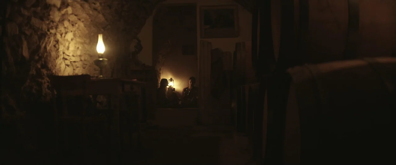 The Only One (2021) movie still: wide — An underground wine cellar with rough stone walls and multiple large wooden barrels. Two people sit together…