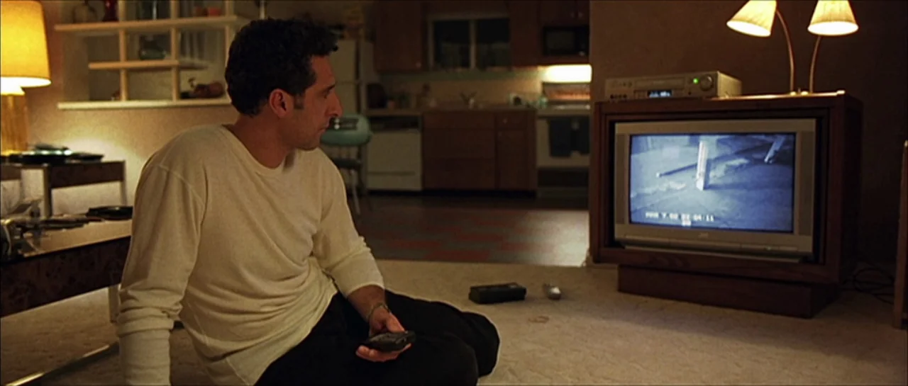 Fear X (2003) movie still: medium — A carpeted room with beige walls and a view into a dimly lit kitchen. A man sits on the floor watching a grainy…