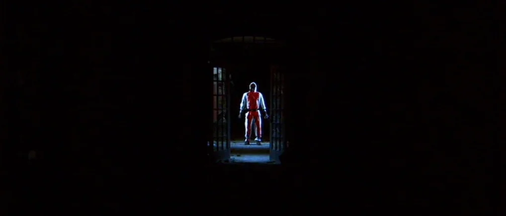 Session 9 (2001) movie still: wide — A dark space features a lone figure framed within an open rectangular doorway. The individual stands perfectly…