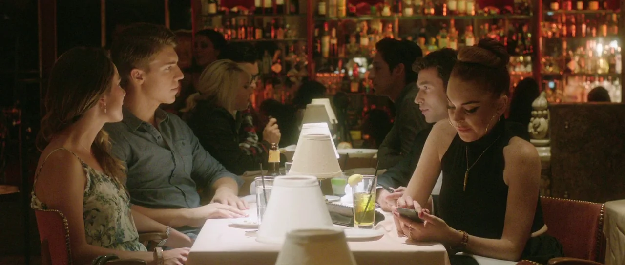 The Canyons (2013) movie still: medium — A dimly lit restaurant booth with a white tablecloth, table lamps, and a background bar. A young woman…