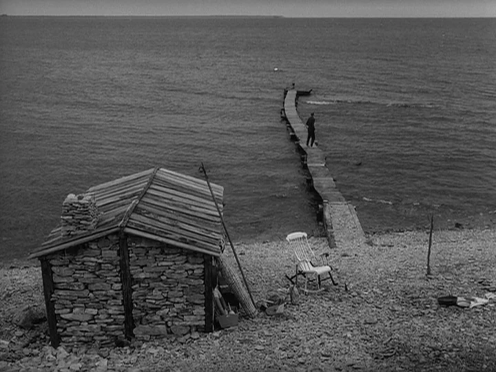 Through A Glass Darkly (1961) movie still: wide — A rocky shoreline features a small stone hut and a wooden pier extending into water. A single man…