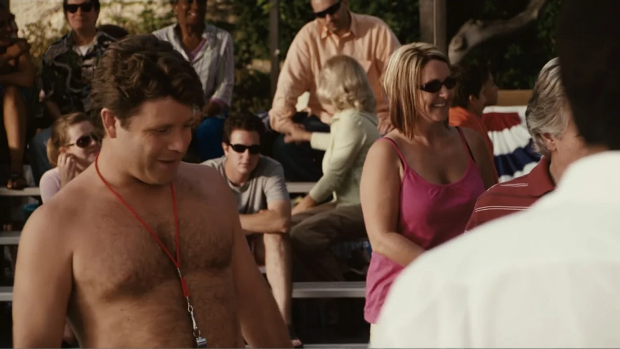 Click (2006) movie still: medium — A group of people sits on metal bleachers outdoors on a sunny day. A shirtless man wearing a whistle lanyard…