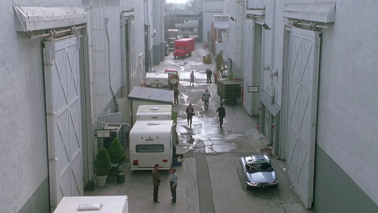Auto Focus movie still: wide — A studio backlot features large industrial buildings flanking a paved pathway with parked trailers. Several people…