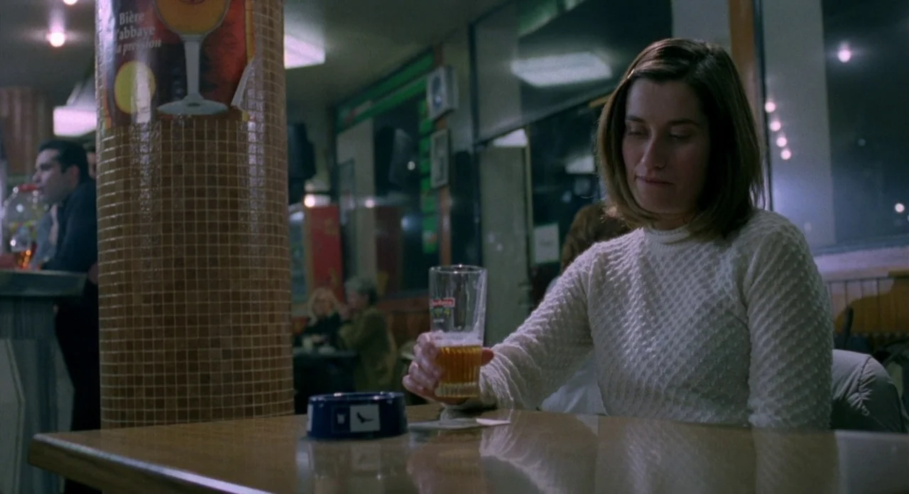 Read My Lips (2001) movie still: medium — A dimly lit tavern with a tiled pillar and scattered tables in the background. A woman sits alone at a…