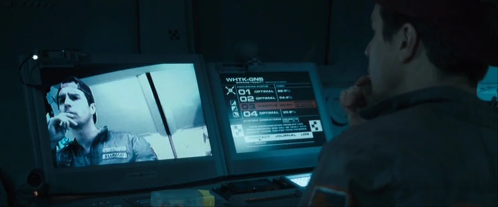 Moon (2009) movie still: medium — A dark, cramped control interface panel illuminates a man's face in cool blue light. The monitor displays a live…