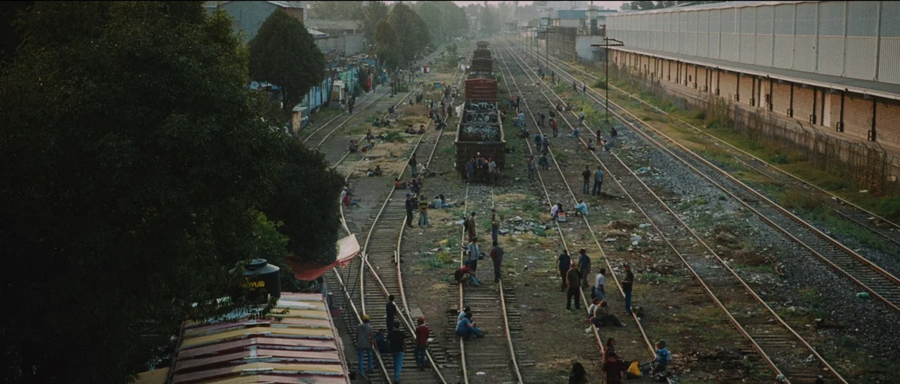 Sin Nombre (2009) movie still: establishing — A expansive industrial railway yard stretches into the distance with multiple converging metal tracks.…