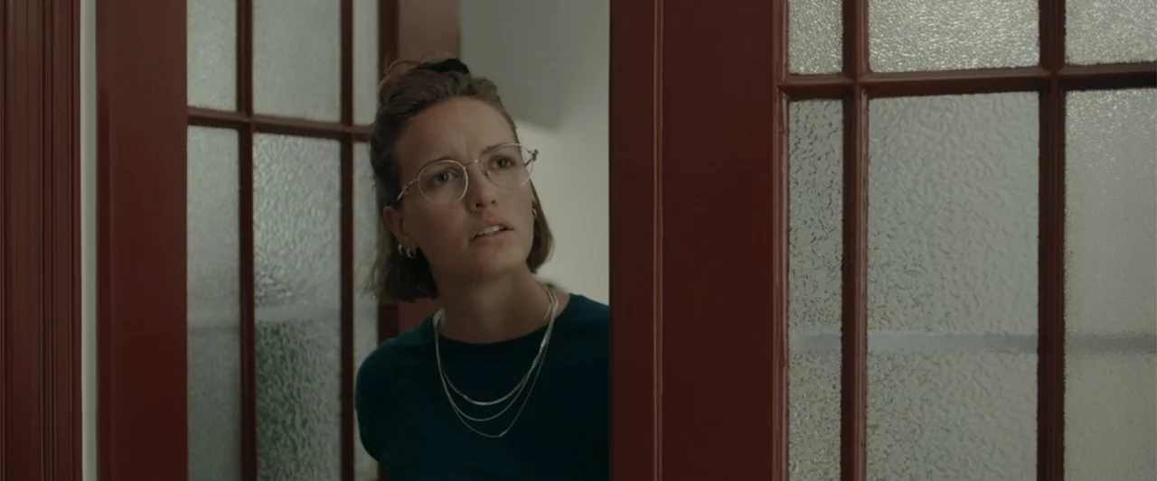 Peacock (2010) movie still: medium — A woman stands in a doorway framed by dark red wood and textured glass panes. The woman looks toward the right…