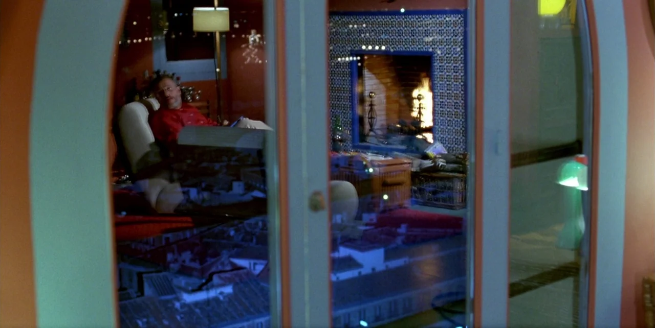 The Flower of My Secret (1995) movie still: medium — An interior space viewed through a glass door reflects a cityscape and the room. A man sits in…