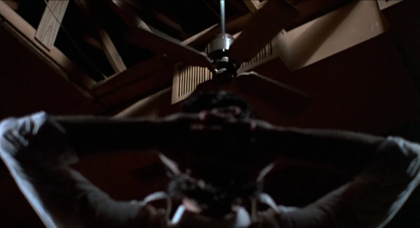 Blood Simple (1984) movie still: medium — A dimly lit room features a wooden ceiling with exposed rafters and a spinning fan. A man lies on his back…